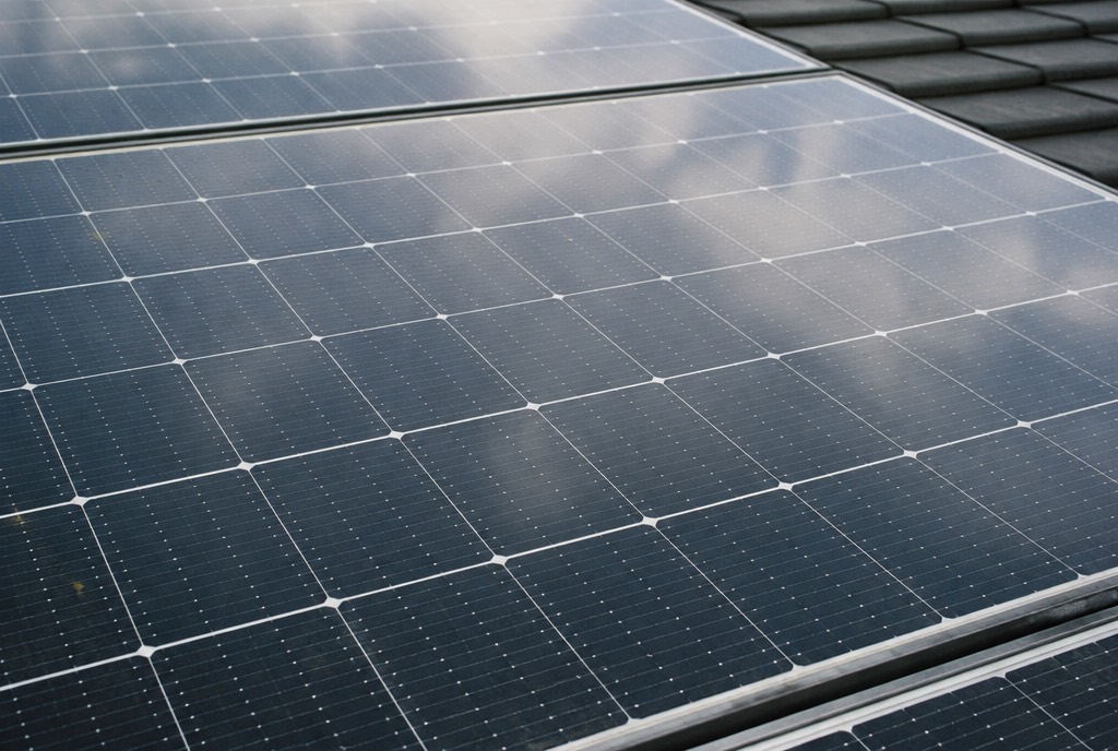 Close-up van zonnepanelen op een dak, waarin een bewolkte lucht wordt weerspiegeld.