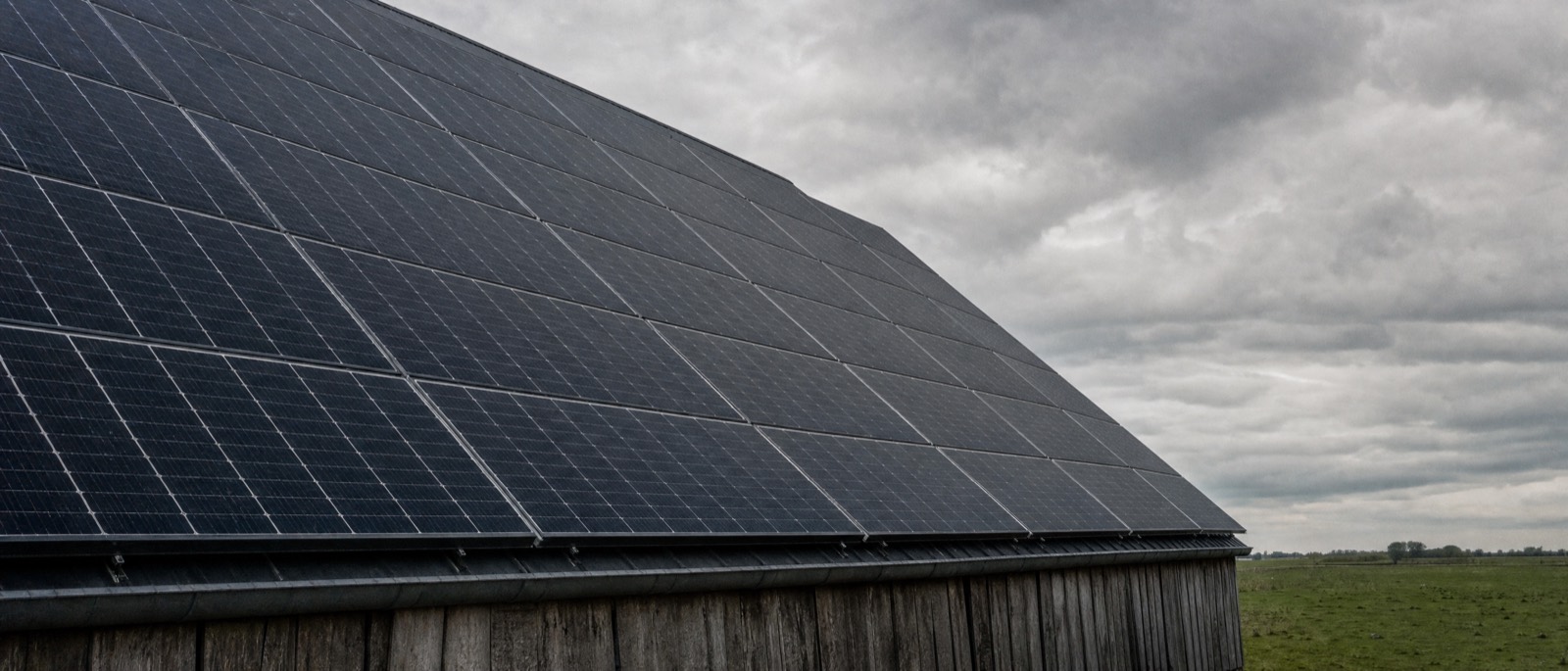 Een groot schuurdak volledig bedekt met donkere zonnepanelen, van onderaf gezien onder een zware bewolkte hemel.